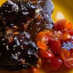 Photo shows chicken with a lustrous dark coating on a plate with sliced cherry tomatoes