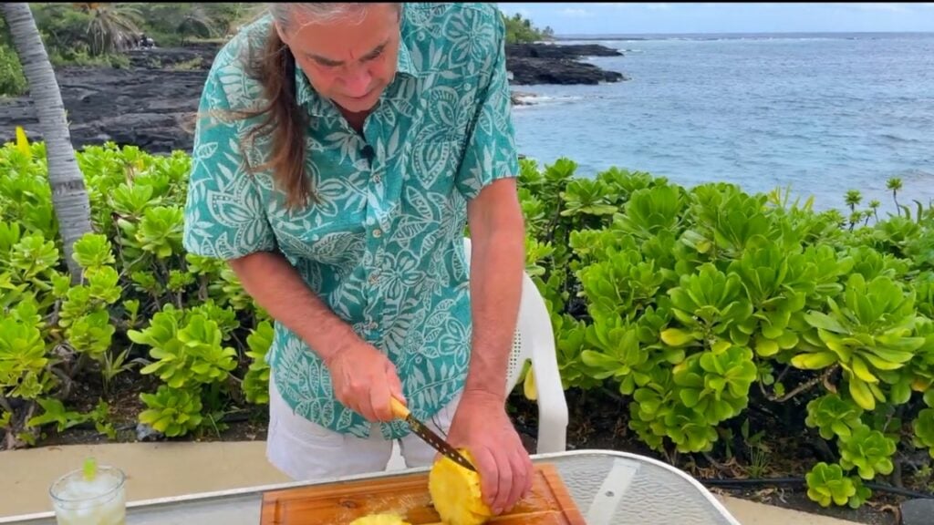 Photo shows cutting a pineapple in Hawaii with greenery and a beautiful blue ocean behind