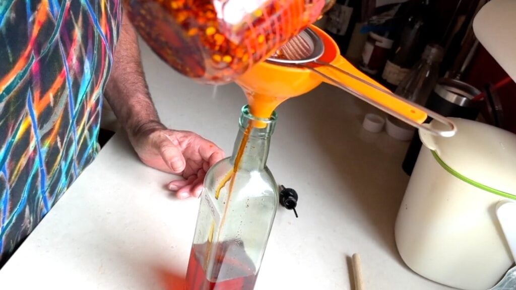 Photo shows chile oil being poured from a measuring cup into a strainer set in a funnel in the oil bottle