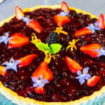 Photo shows a blueberry cheesecake tart decorated with strawberry slices, mint and edible flowers